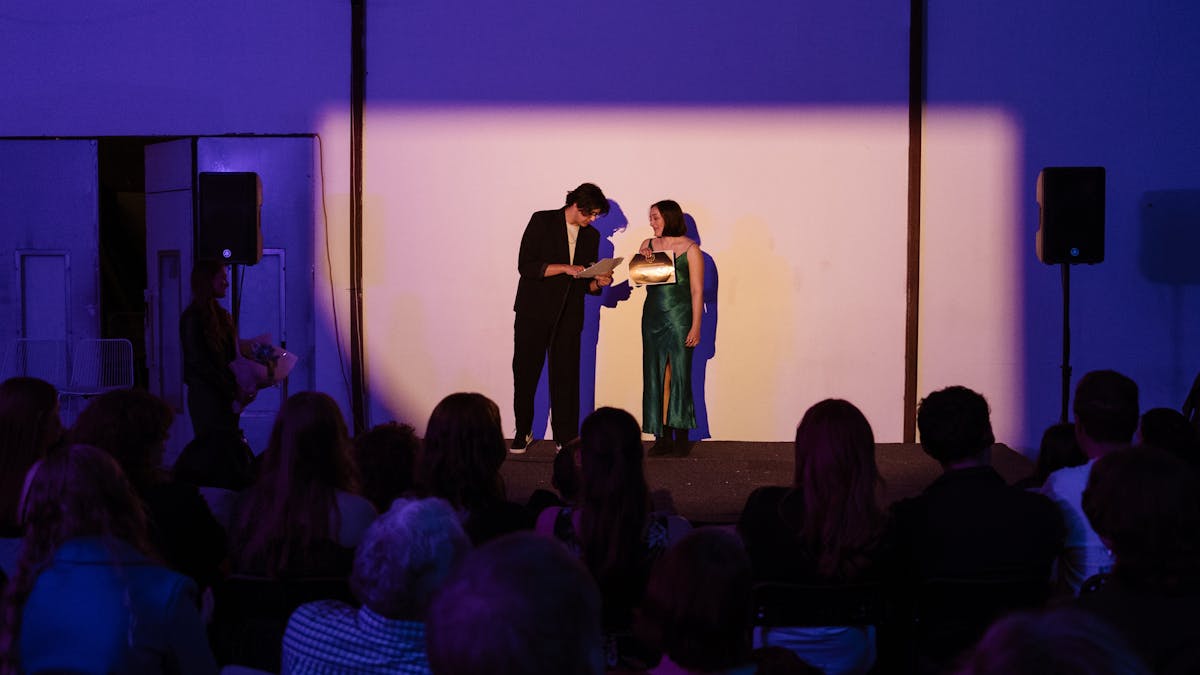 Two people stand on a stage, reading from a paper under a spotlight. An audience watches attentively