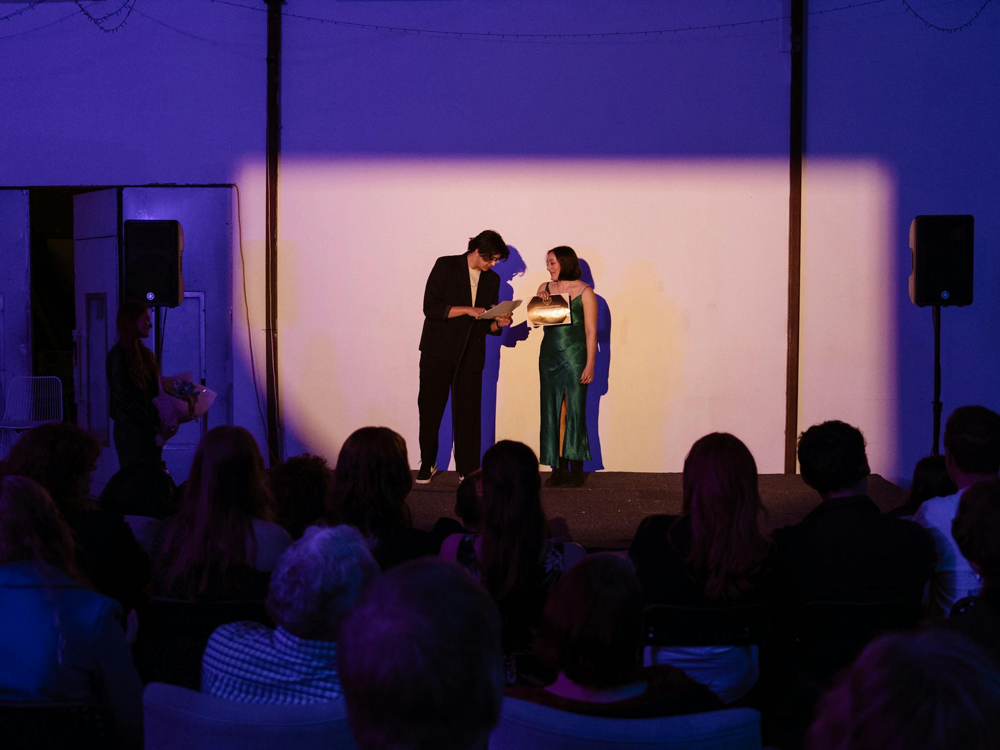 Two people stand on a stage, reading from a paper under a spotlight. An audience watches attentively Two people stand on a stage, reading from a paper under a spotlight. An audience watches attentively