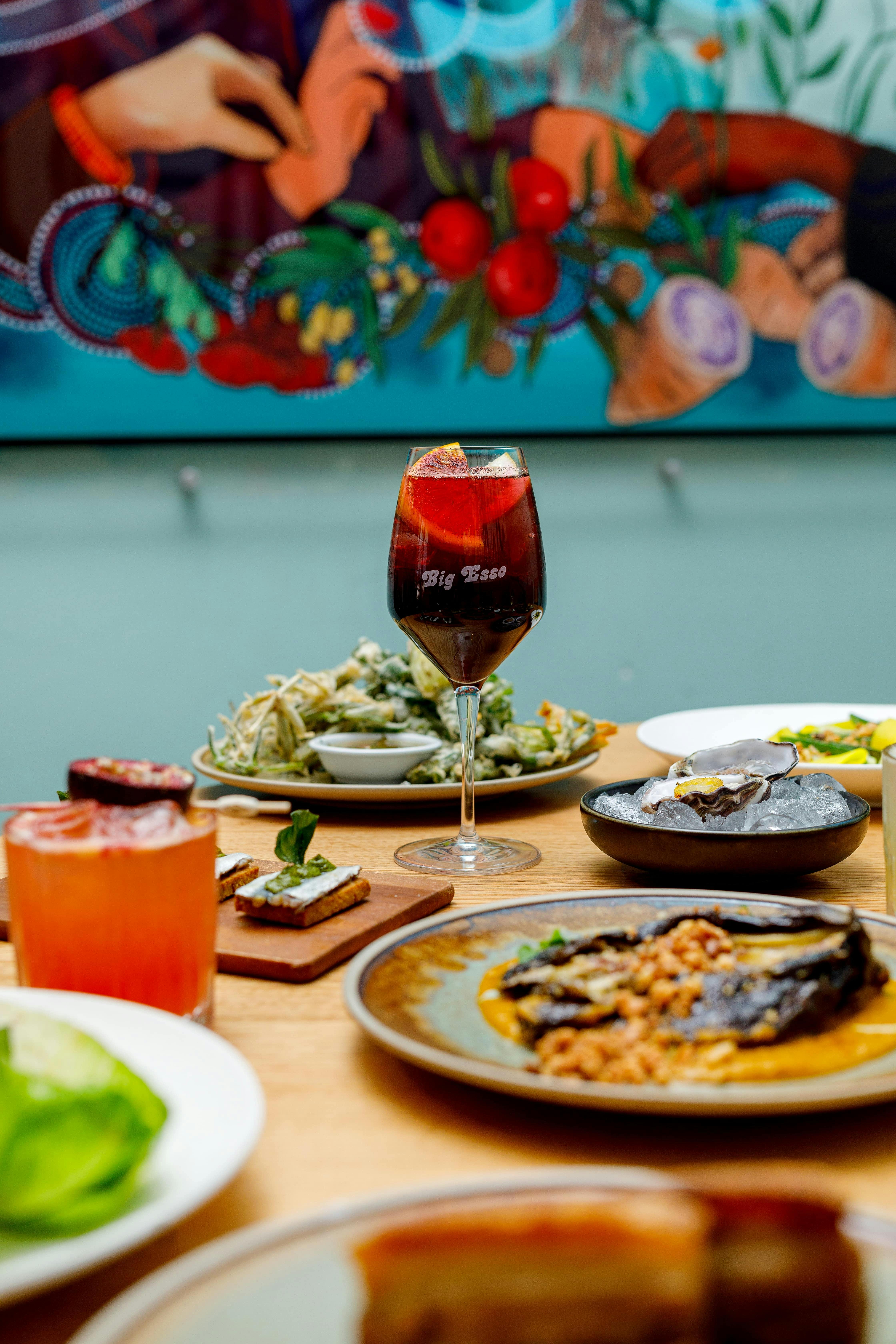 Wine glass with sangria reading "big esso" surrounded by plates of food. Blue mural in background.
