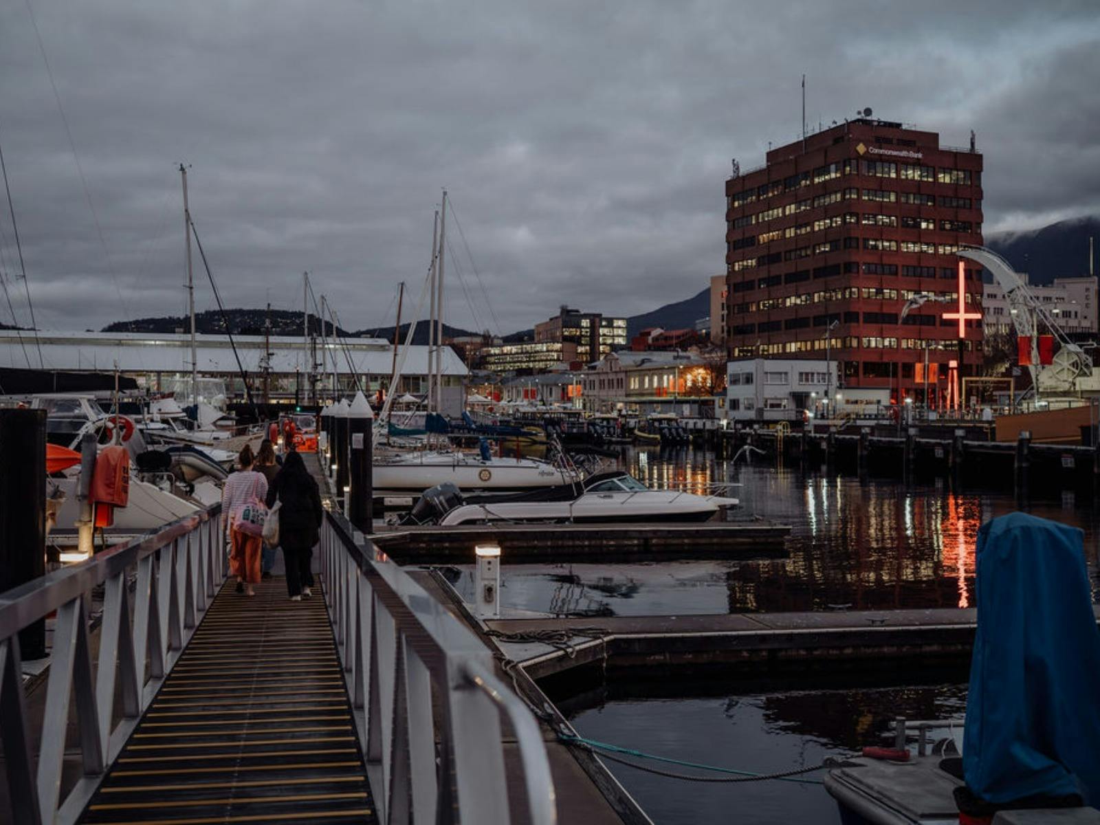 People walk down a pier gangway at dusk toward a harbor and a glowing red cross.