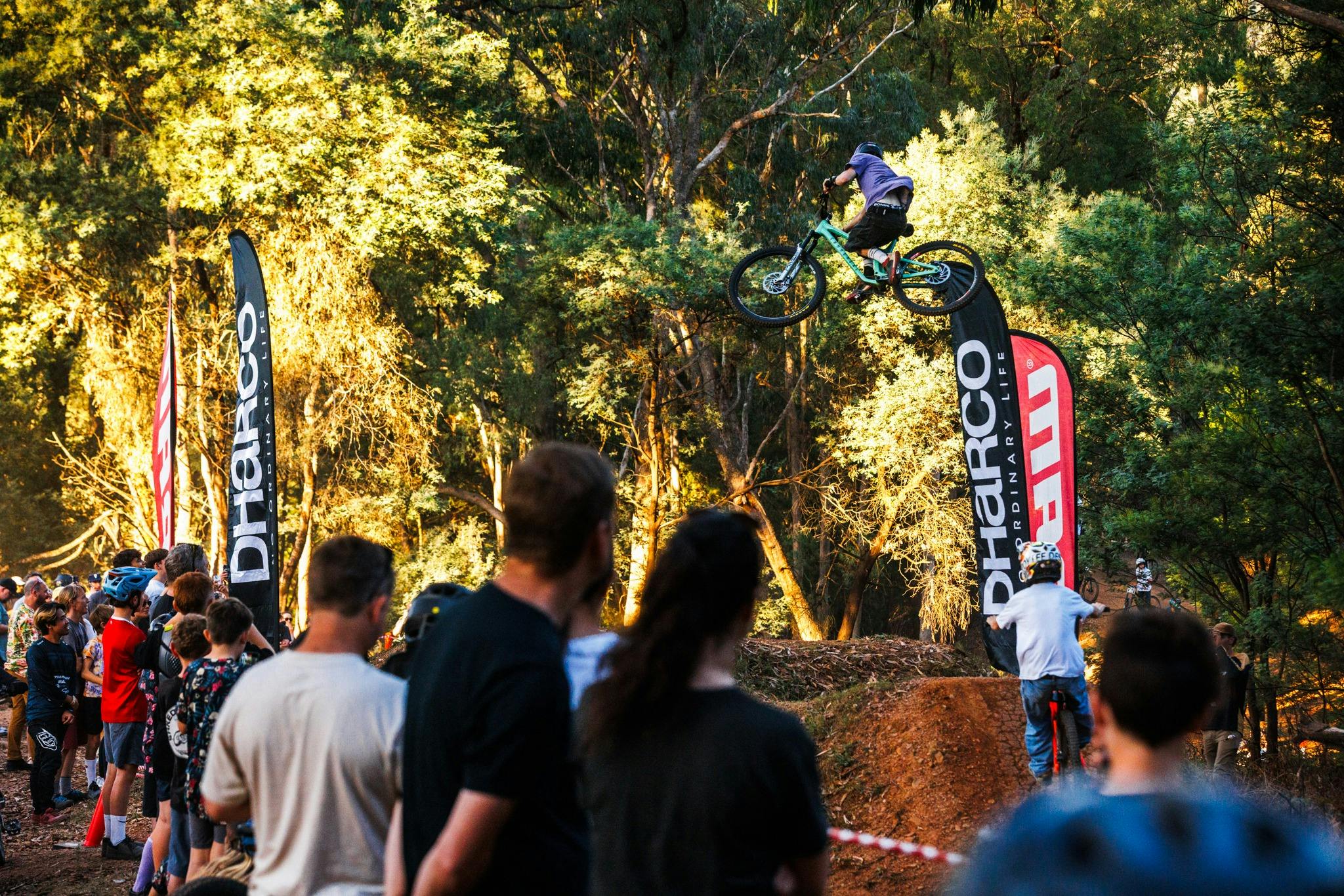 Rider jumping a bicycle between dirt jump surrounded by crowd and sponsor flags at Mystic Bike Park