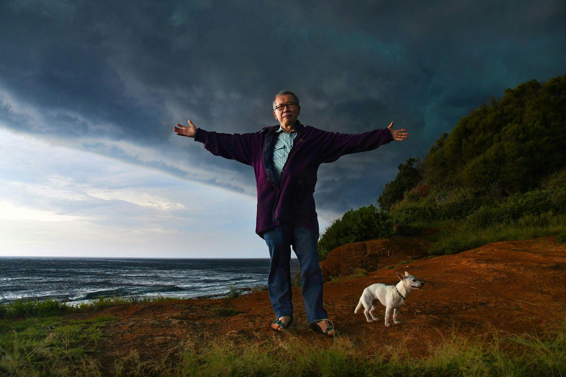 A person stands on a hilltop, arms outstretched, accompanied by a small white dog