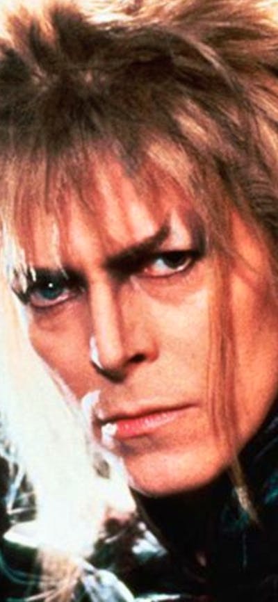 A man with wild blonde hair holding something in a black-gloved hand staring at the camera