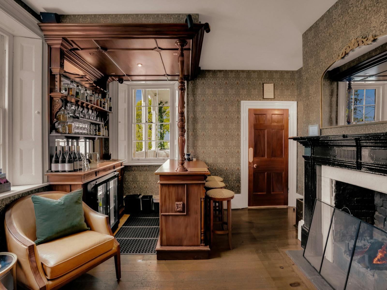 Small wallpapered room with wooden bar stocked with alcohol, open fire place and chairs