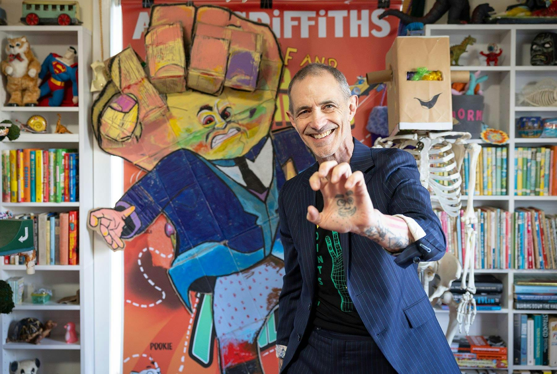 A man in a striped suit smiles and holds up his hand, which has a face tattooed in the palm.