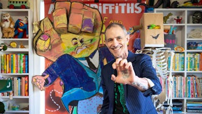 A man in a striped suit smiles and holds up his hand, which has a face tattooed in the palm.