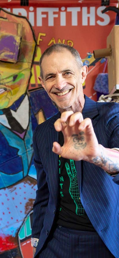 A man in a striped suit smiles and holds up his hand, which has a face tattooed in the palm.
