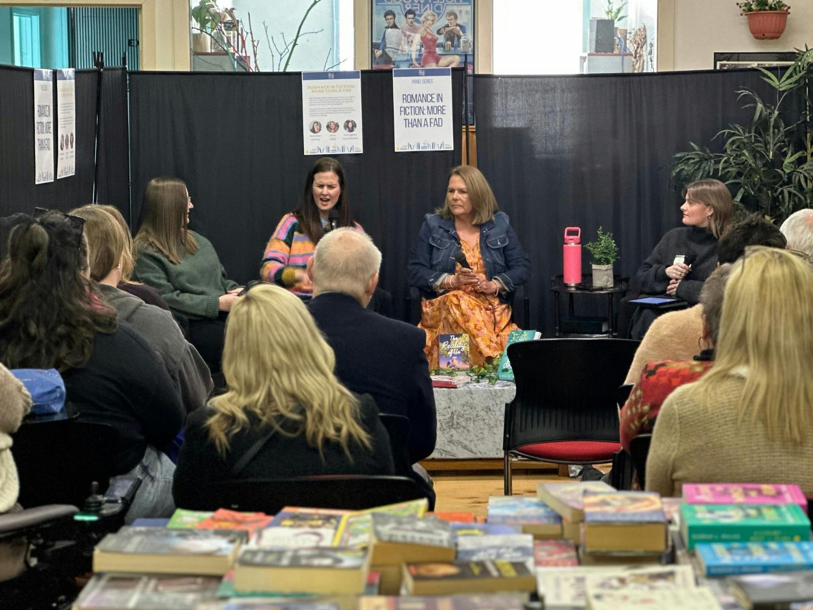 Background features four women talking about romance books, the audience blurred in the foreground.