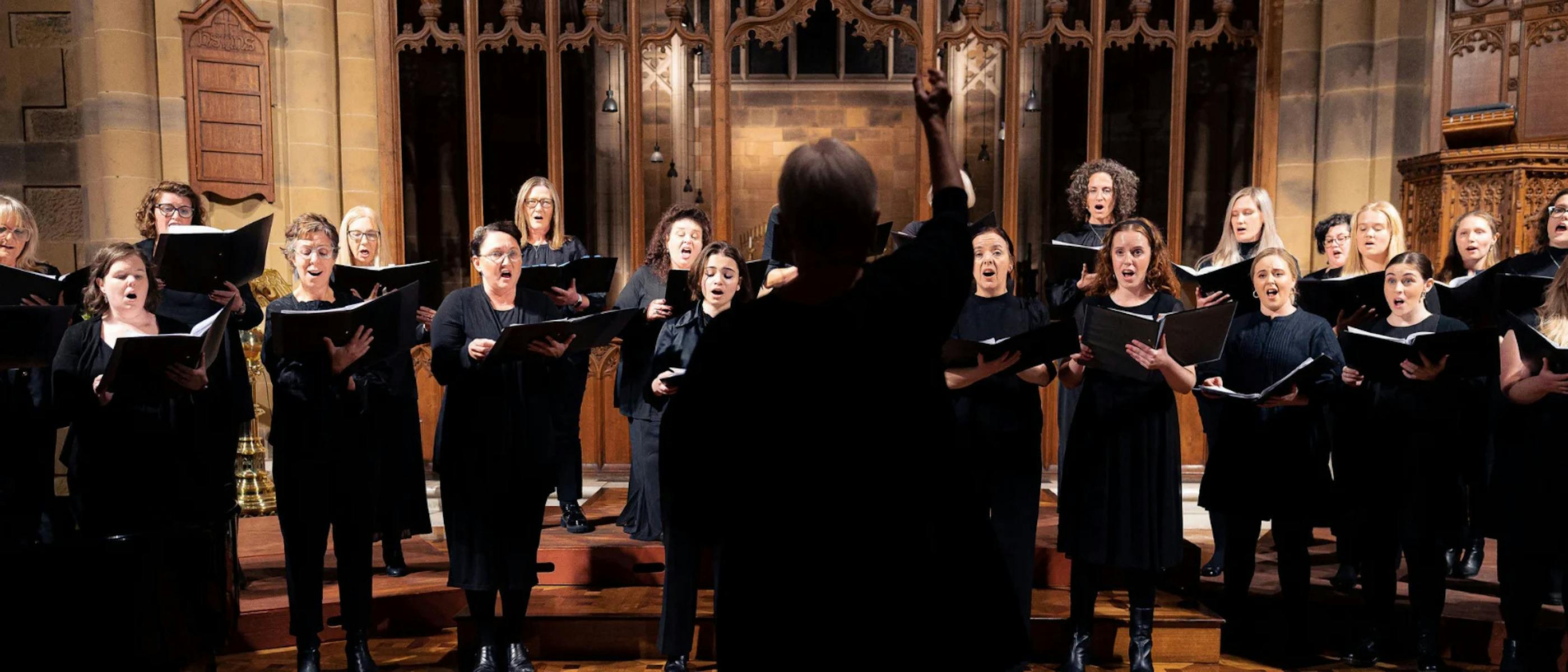 A group of singers wearing black and holding songbooks, being conducted.