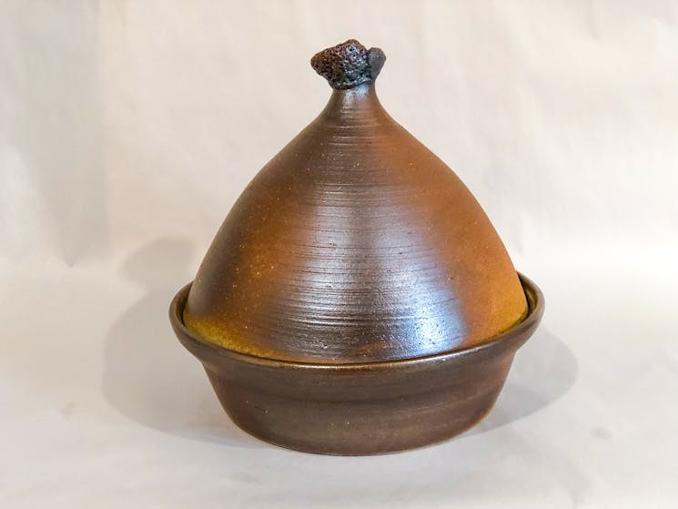 A large dark brown tagine pot, cream background. Shiny surface, handle on top leaning to the left.