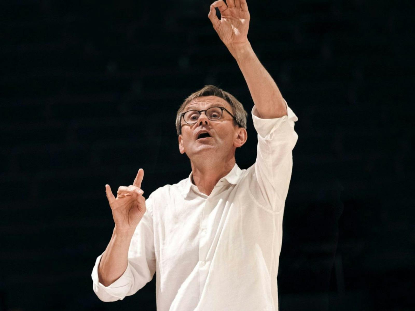 A conductor, wearing a white shirt, arms lifted in the air,  standing against a black backdrop.