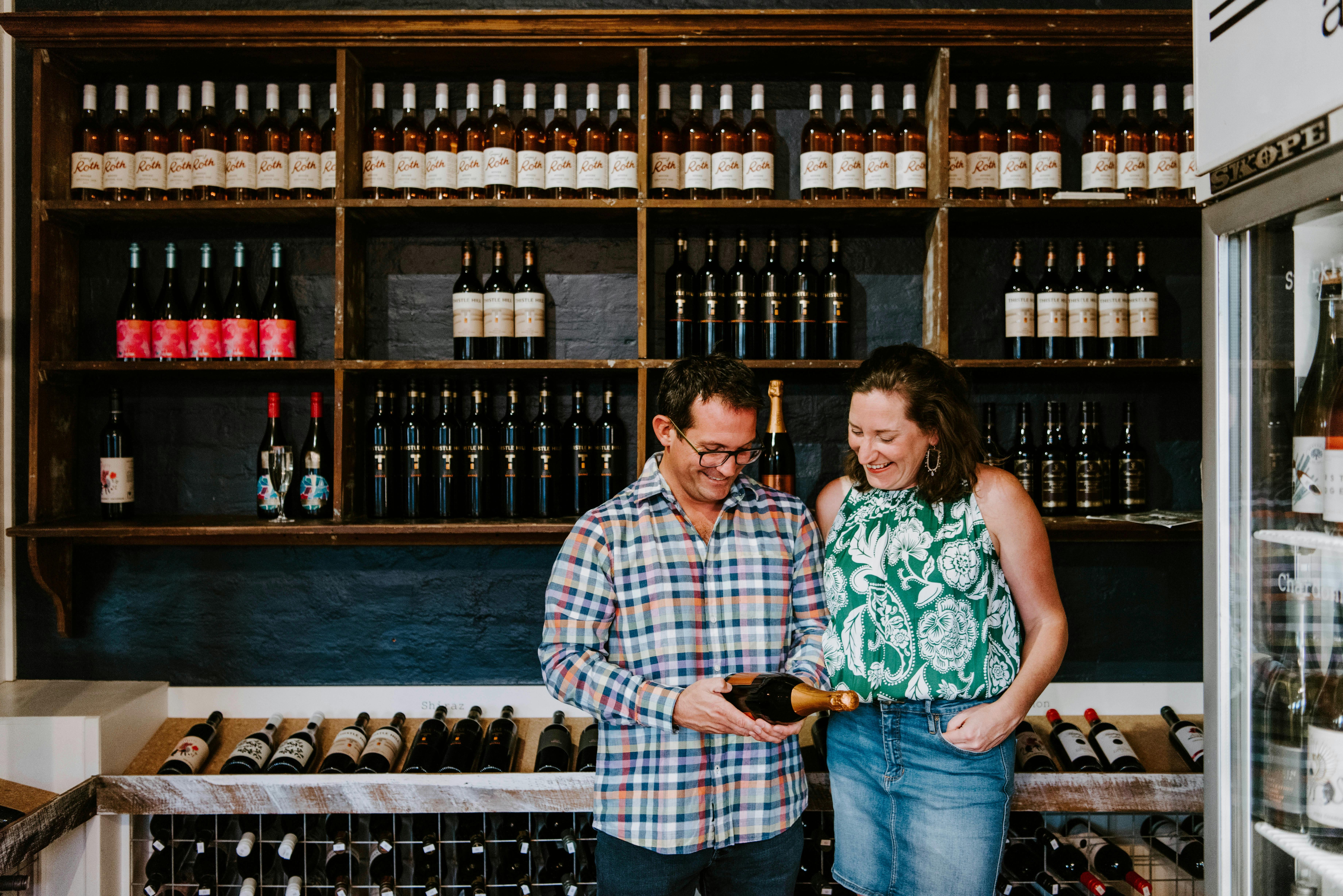 Couple in bottle shop looking at wine bottle, backdropped by shelves of wine
