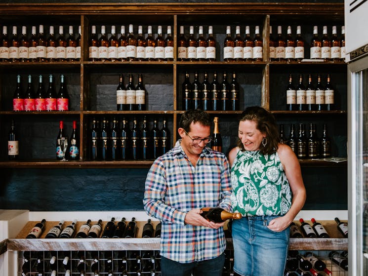 Couple in bottle shop looking at wine bottle, backdropped by shelves of wine