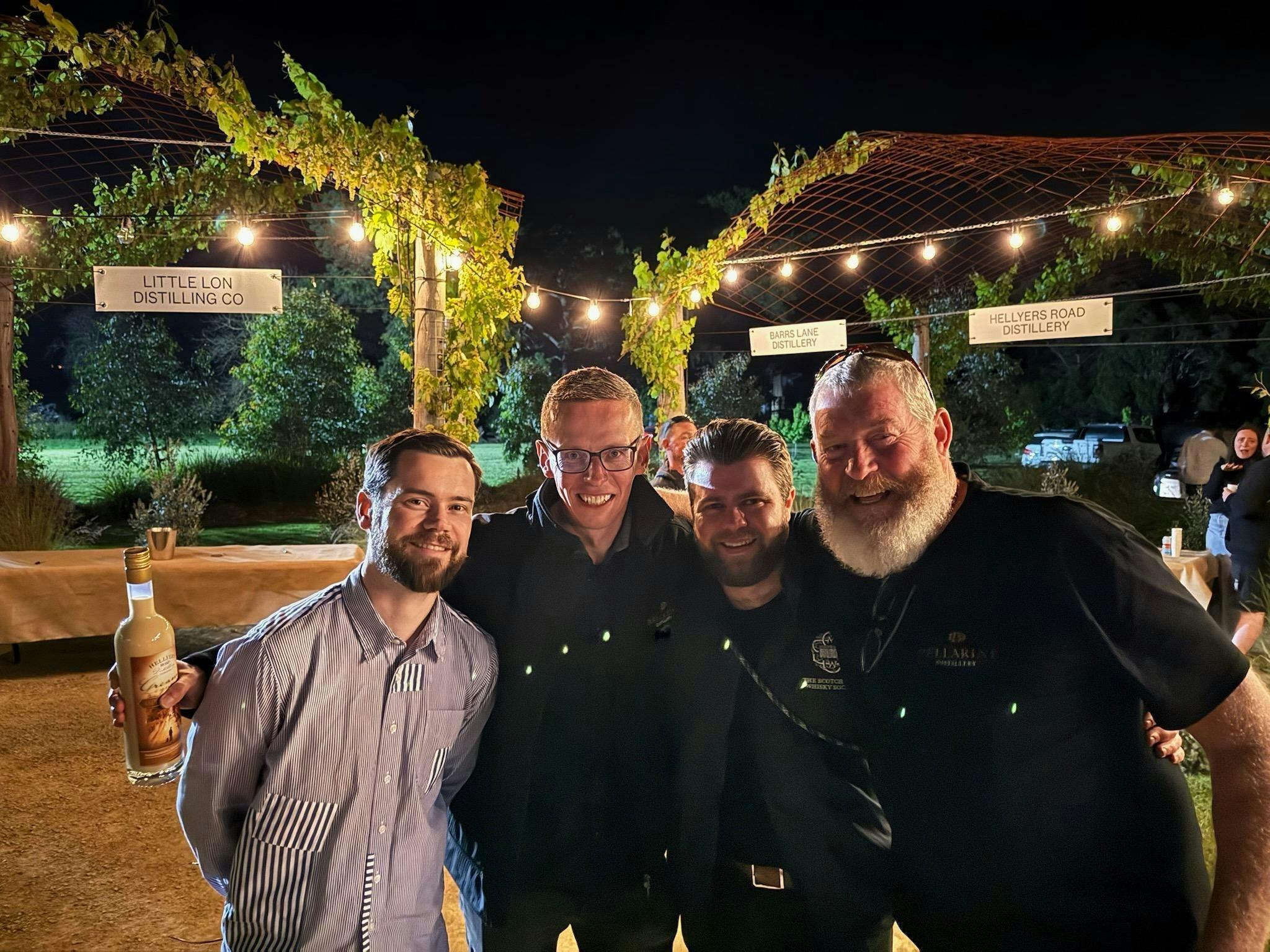 Four Australian whisky makers smiling beneath festoon lights and vine-covered pergolas at Whiskery