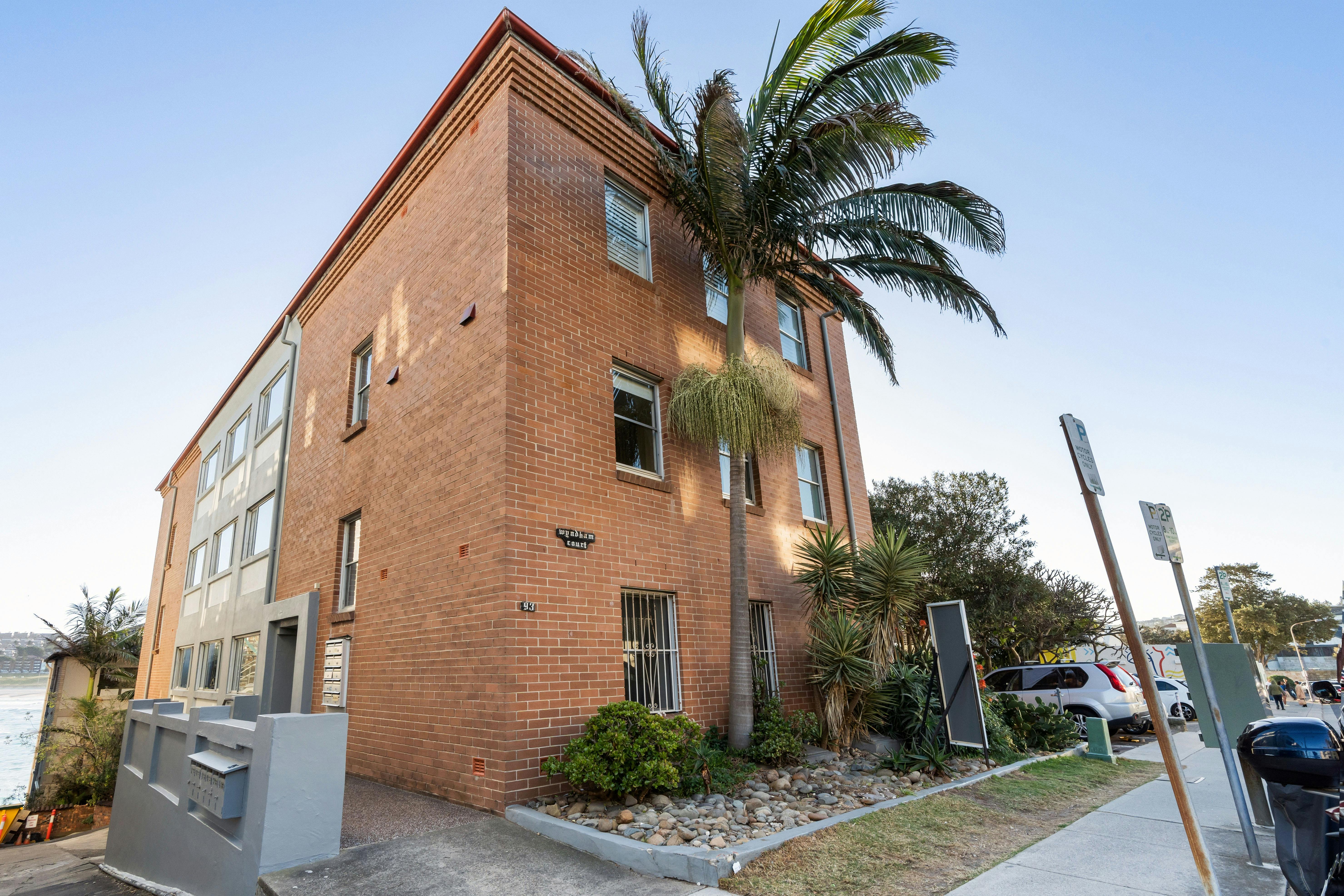 Exterior view of a modern brick apartment building with palm trees and parking nearby.