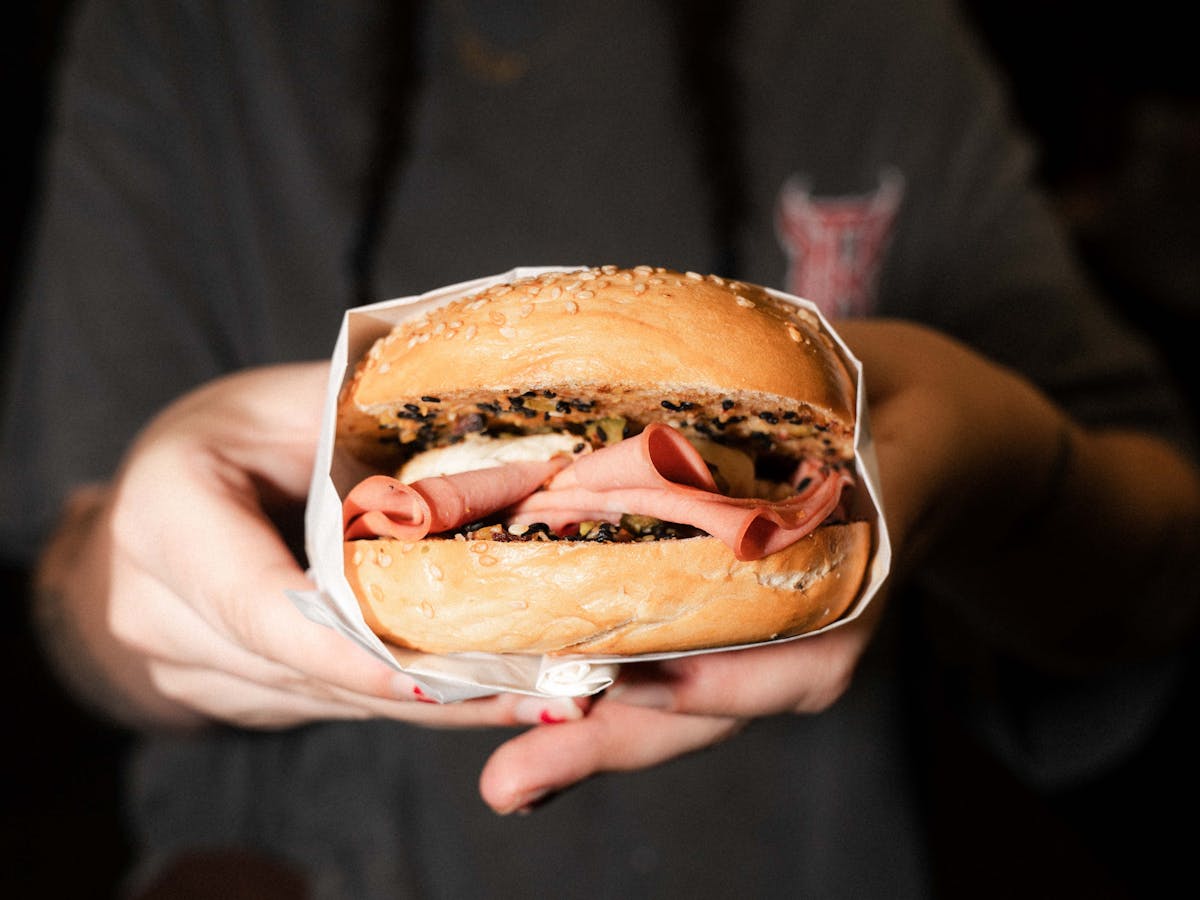 Two hands holding a seeded bagel with mortadella and mozzarella at the focus of the photo