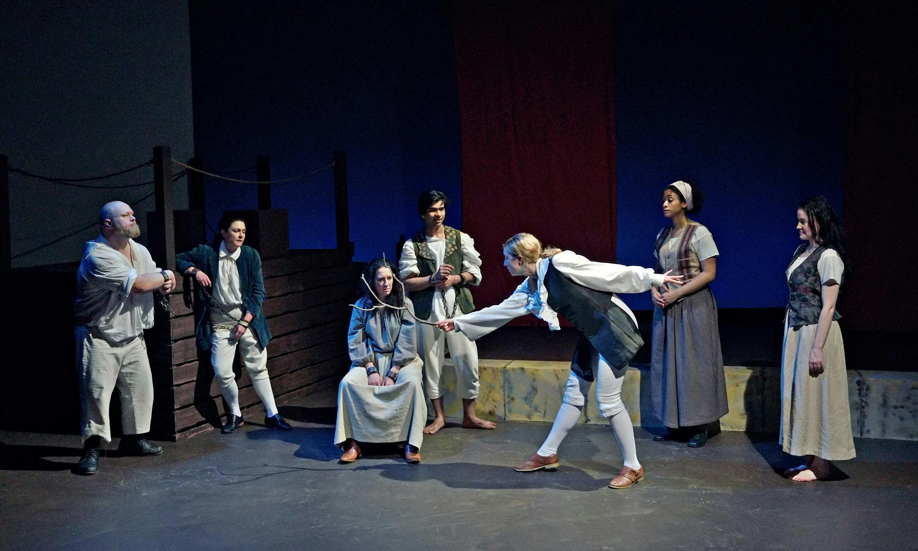 scattered group dressed as convicts - a woman sits centre in shackles being annoited with a stick