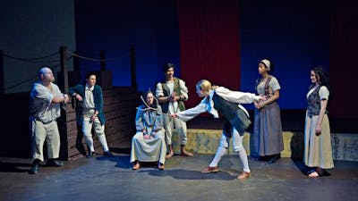 scattered group dressed as convicts - a woman sits centre in shackles being annoited with a stick