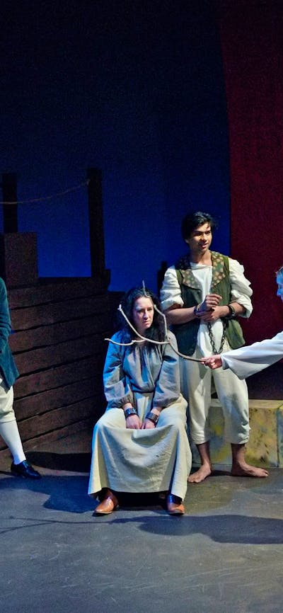 scattered group dressed as convicts - a woman sits centre in shackles being annoited with a stick