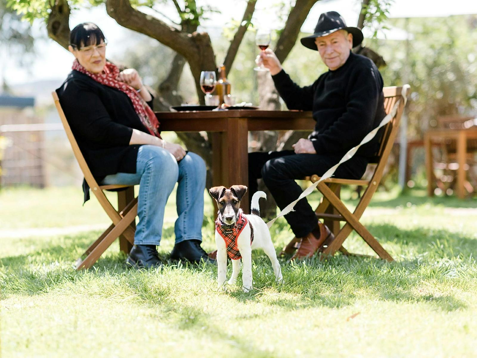 two people at a table drinking wine with their dog standing by the table