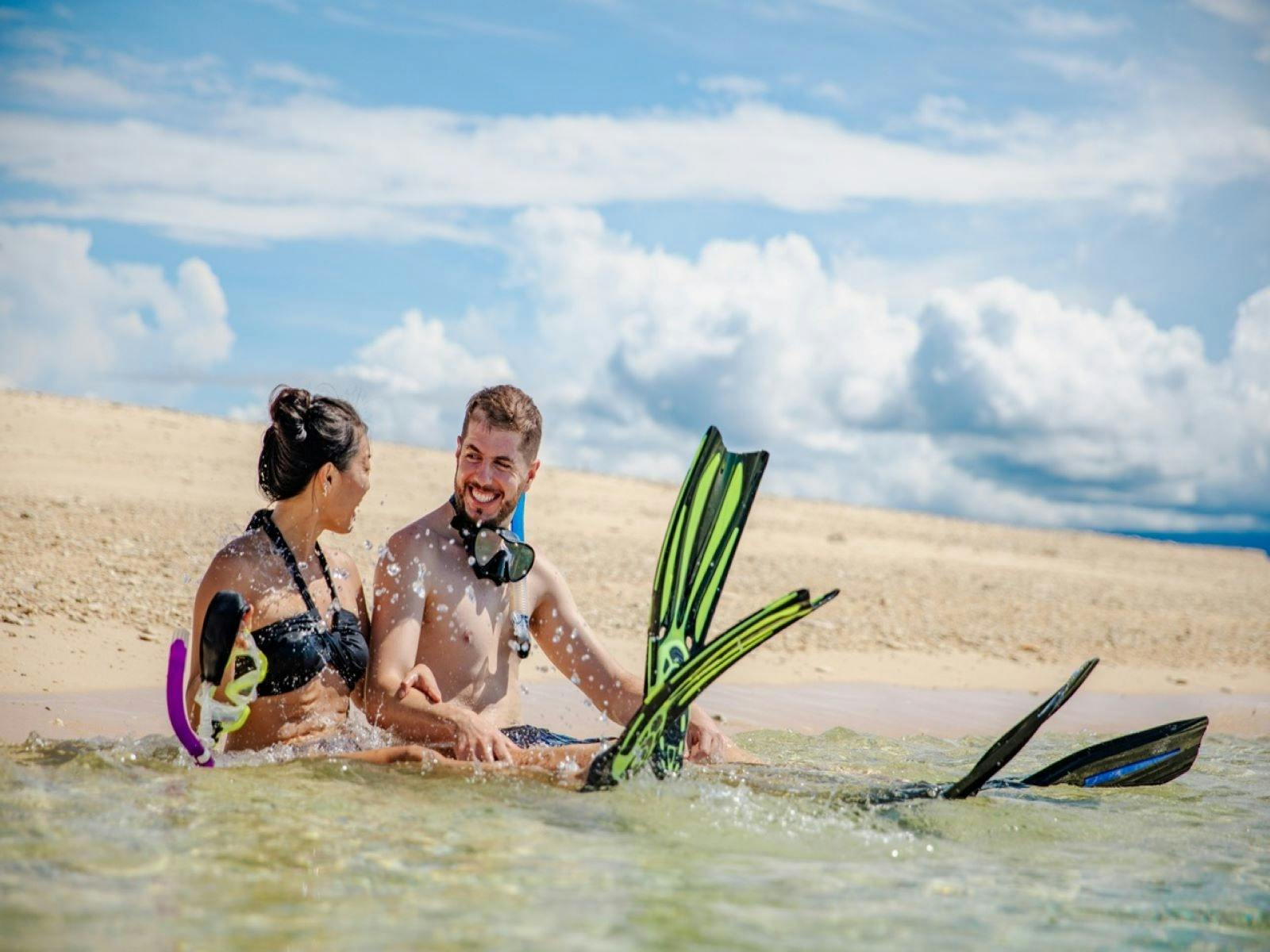 Couple sitting along beach, smiling and putting on snorkel gear