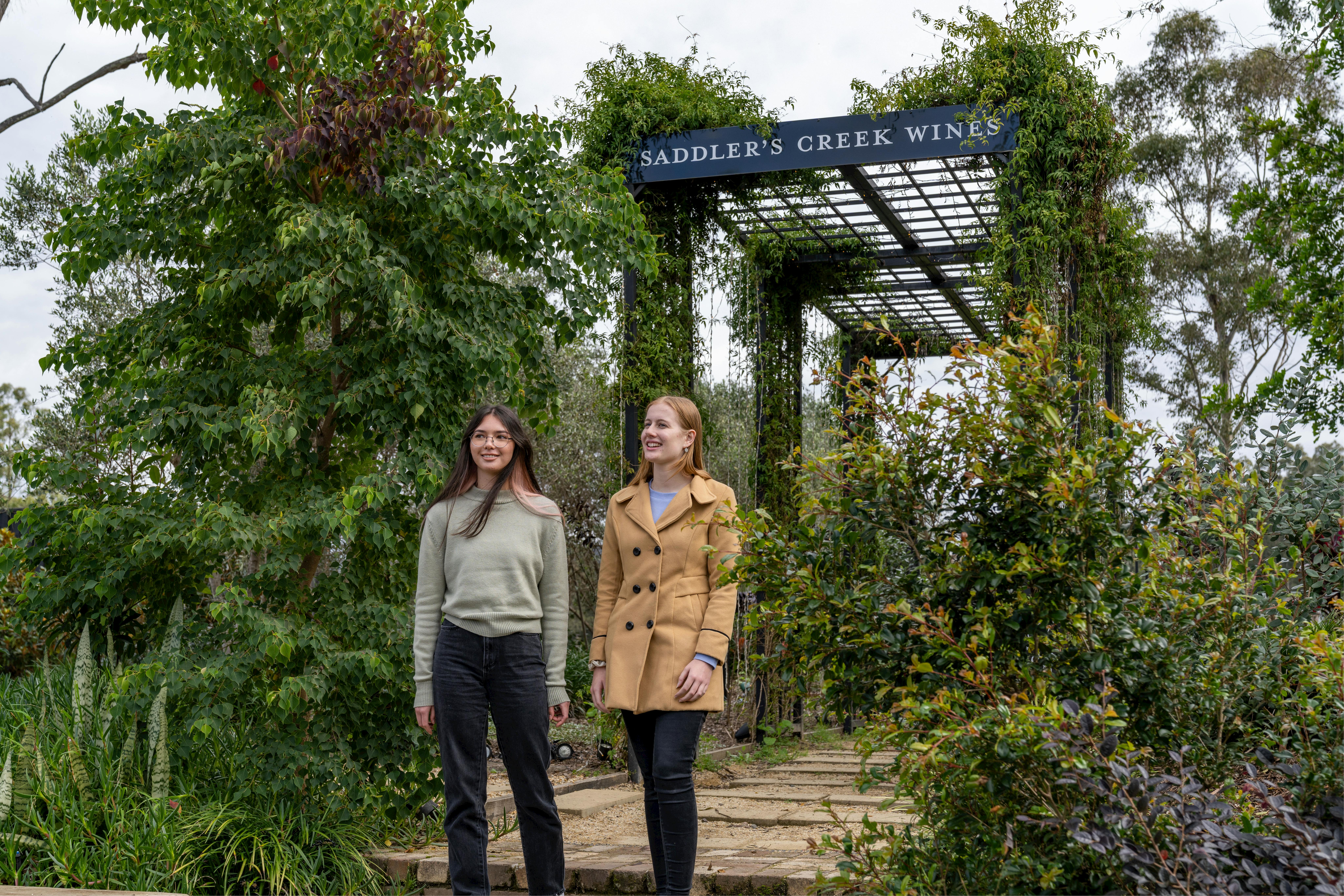Zwei Frauen gehen unter der Eingangslaube von Saddler's Creek Wines spazieren, umgeben von Grün