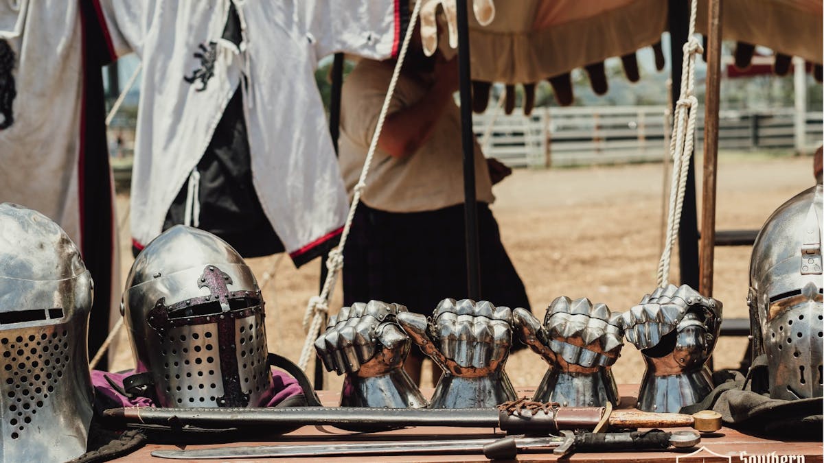 Medieval knight helmets, gauntlets, and swords on display at a festival encampment.