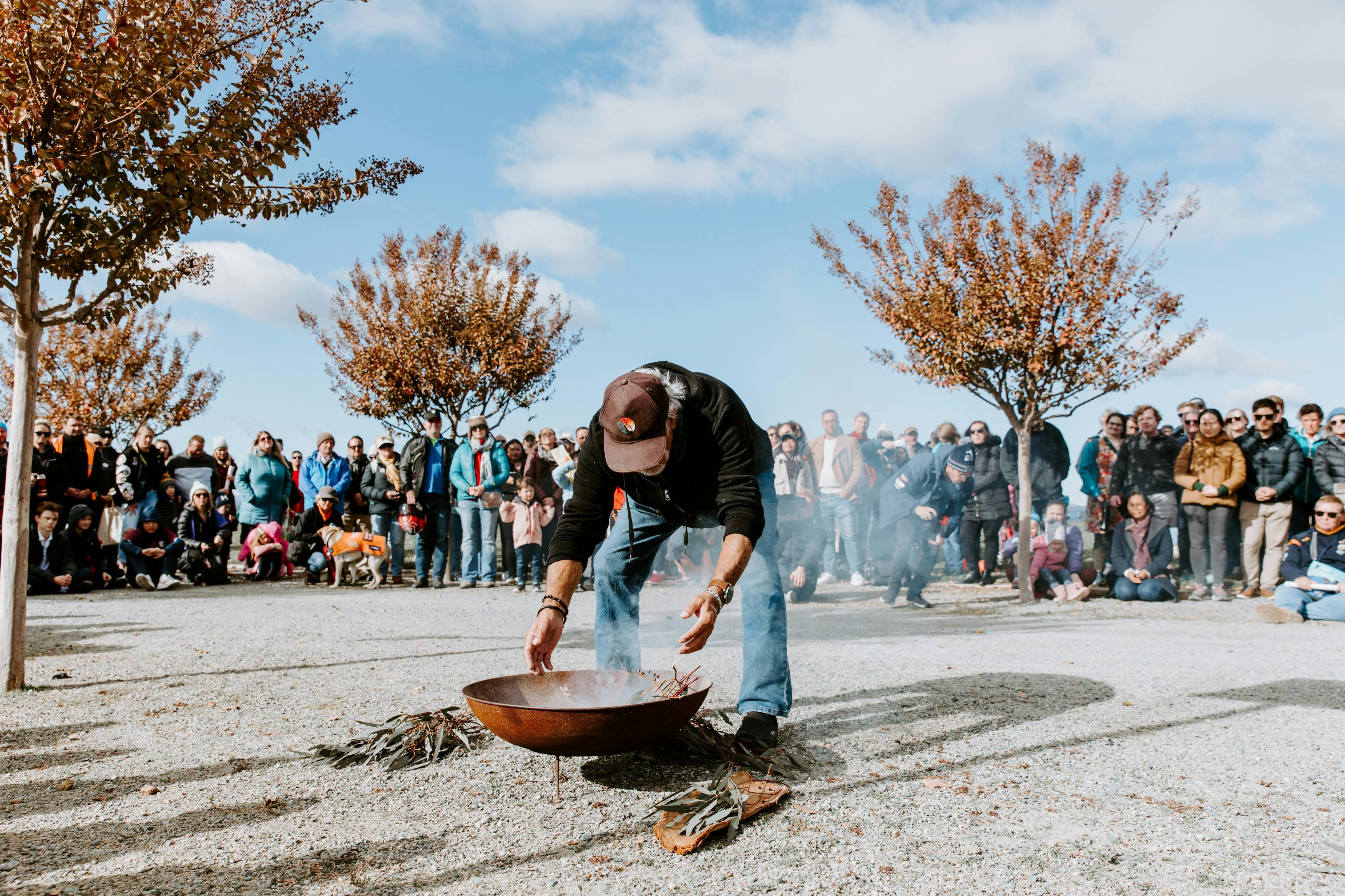 Person tending to a fire pit during a smoking ceremony, surrounded by a large crowd