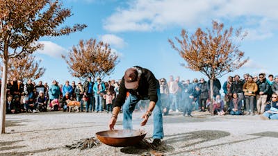 Person tending to a fire pit during a smoking ceremony, surrounded by a large crowd