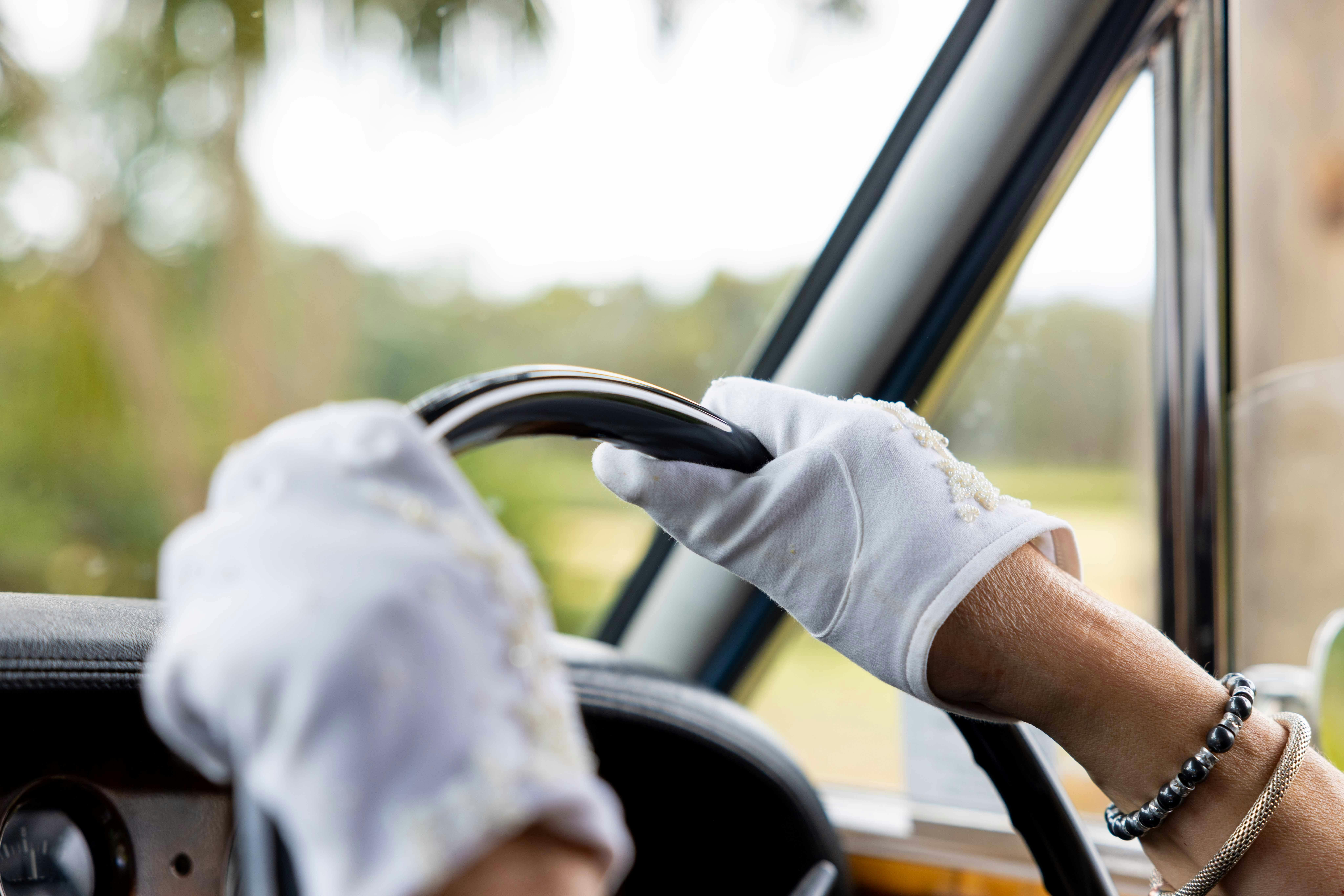 A chauffeur wears white vintage gloves with beaded detail.  Gloved hands steer the Rolls Royce