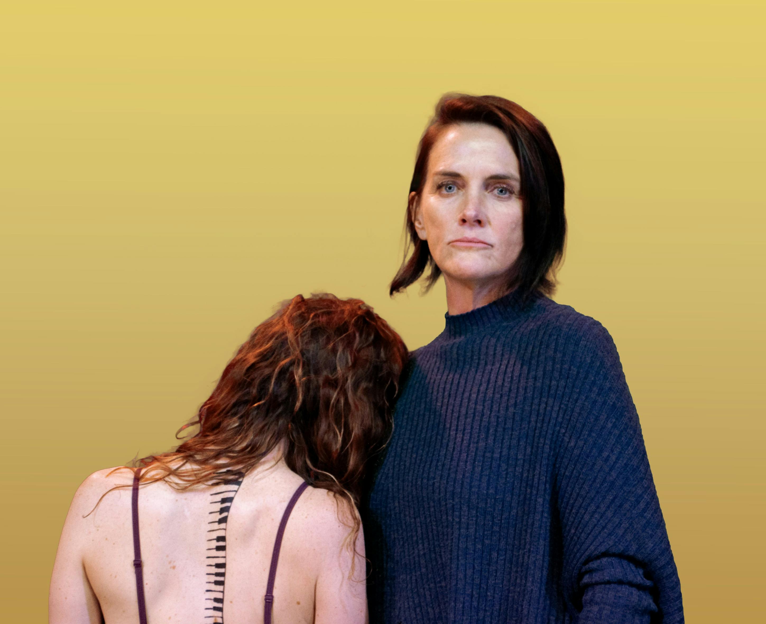 a stoic woman looks straight into the camera while a naked woman leaning on her facing away