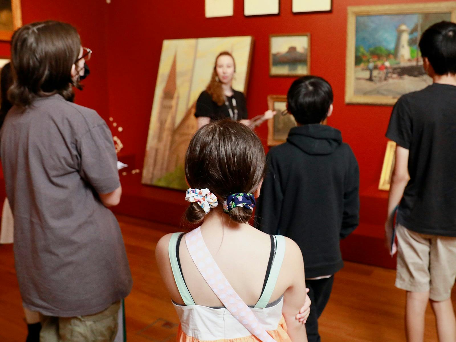A group of people stand listening to a Museum tour guide in a red-walled gallery space.