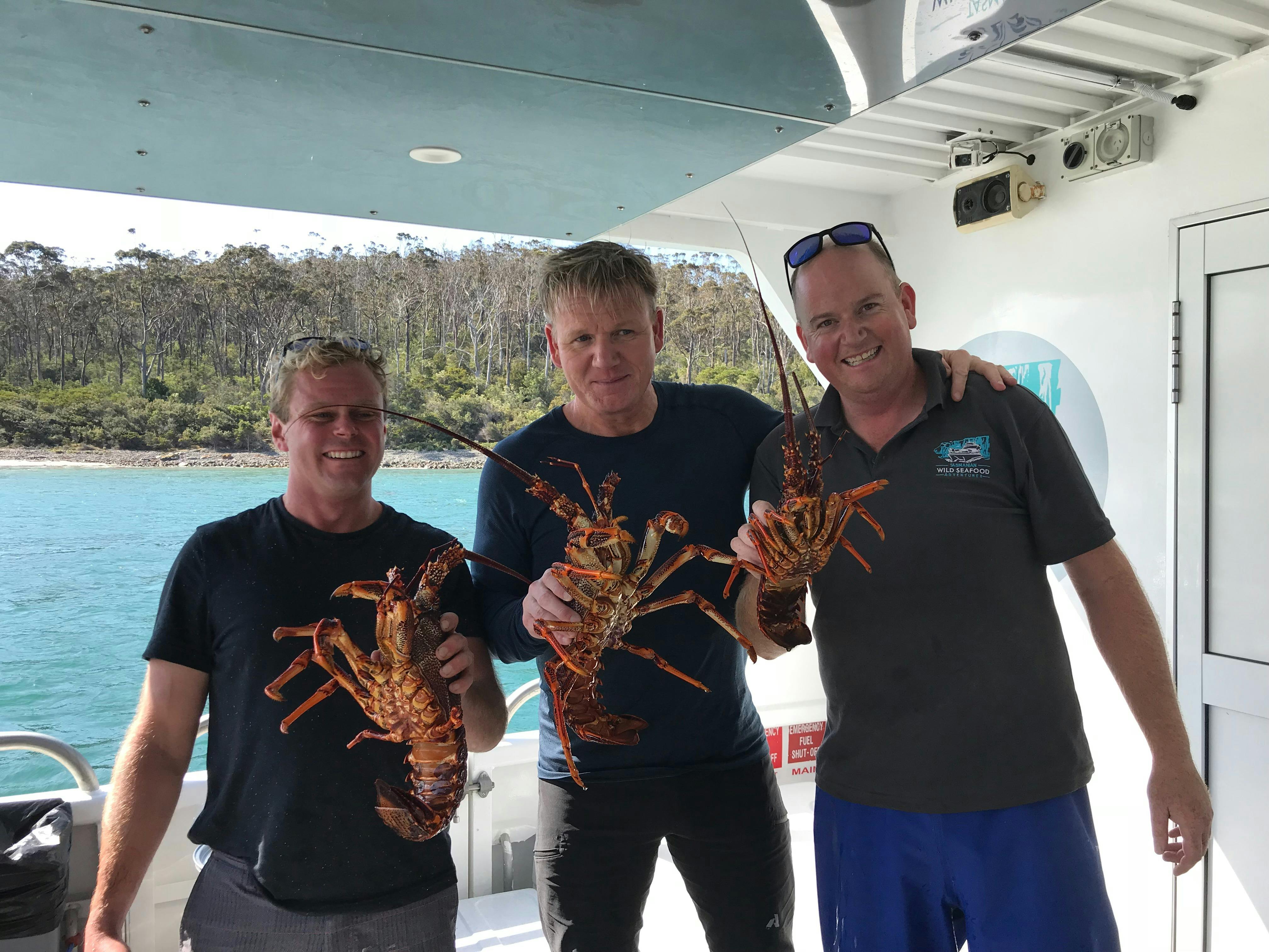 Three men are on a vessel with the water in the background while they each hold a rock lobster