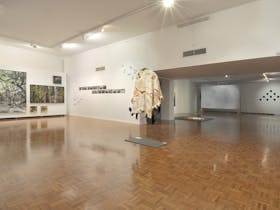 Open gallery space with indigenous animal skin cape hanging from ceiling and forest scene paintings