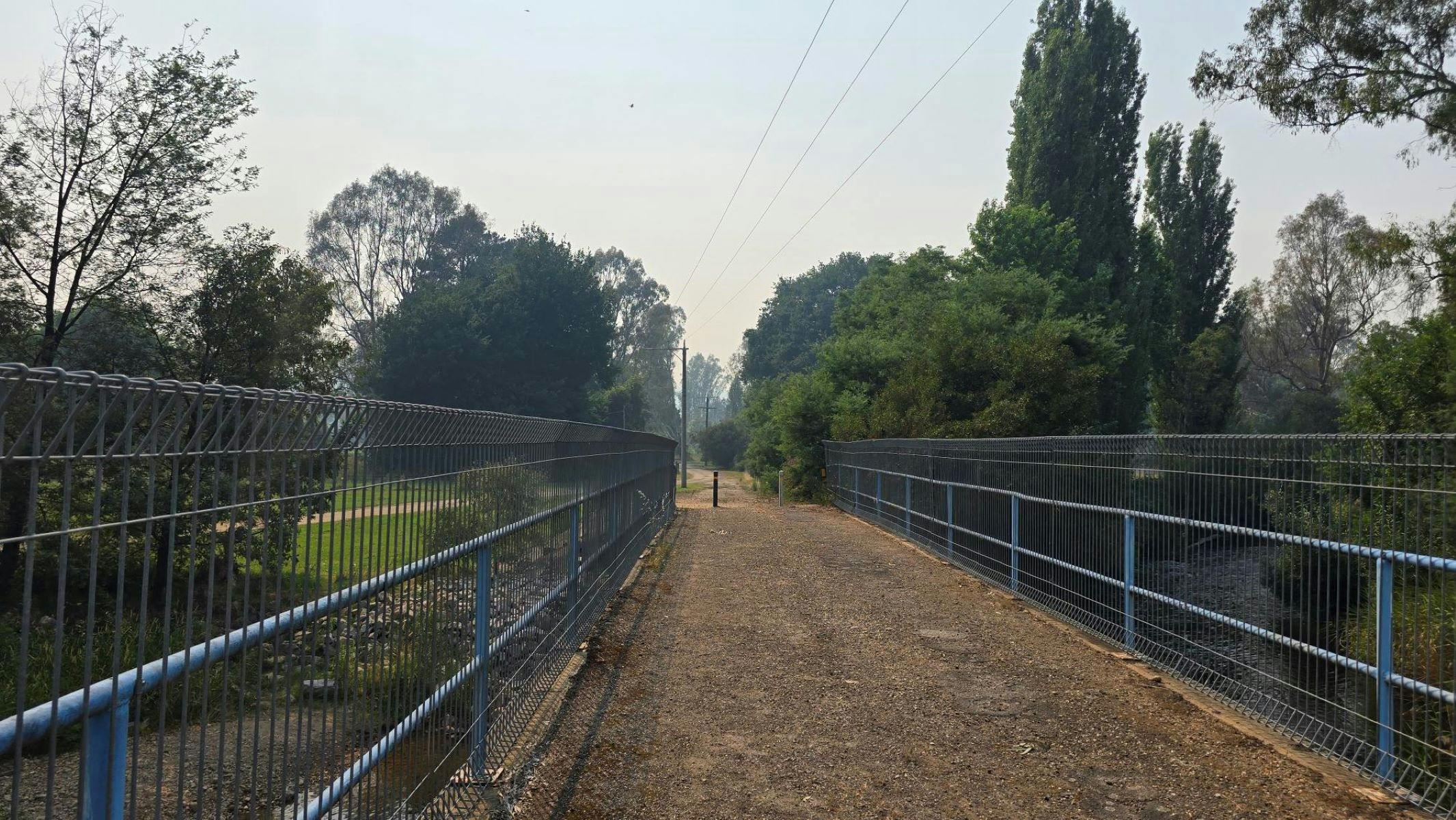 Gravel path going over a bridge, lined with fencing. Trees in the background