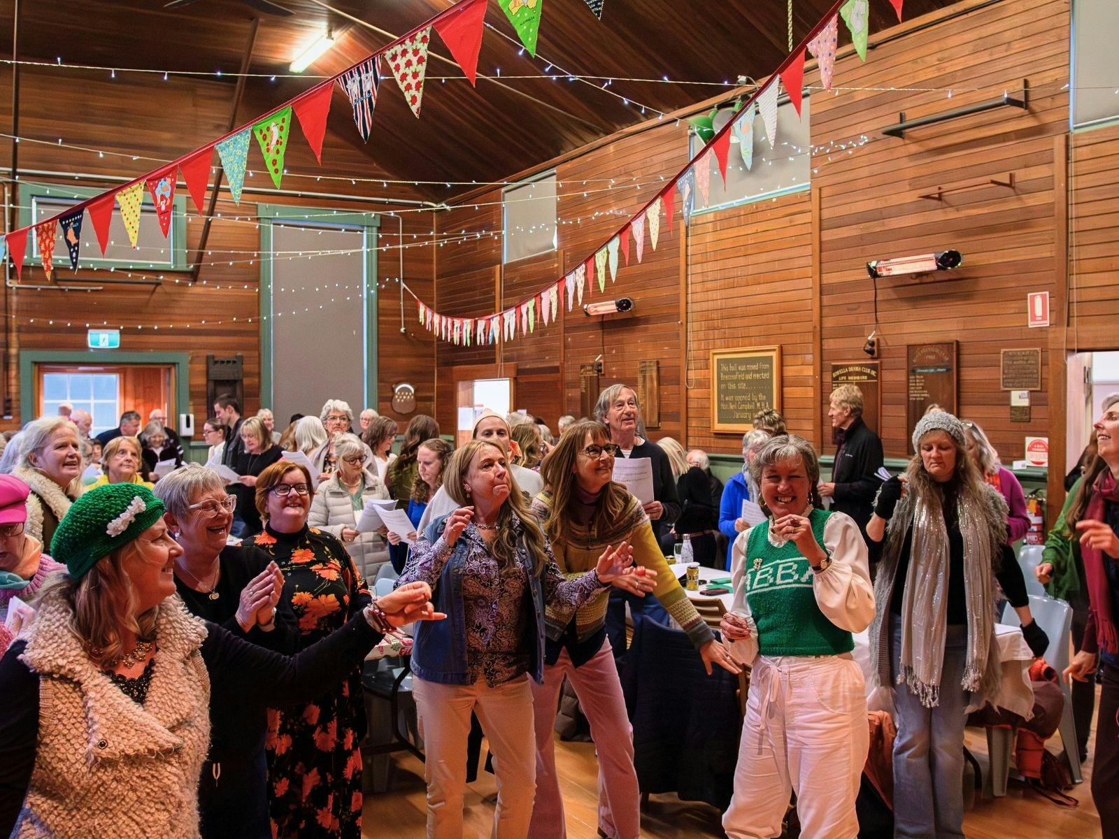 Adults singing, clapping, and moving in a wood-paneled hall with colorful bunting