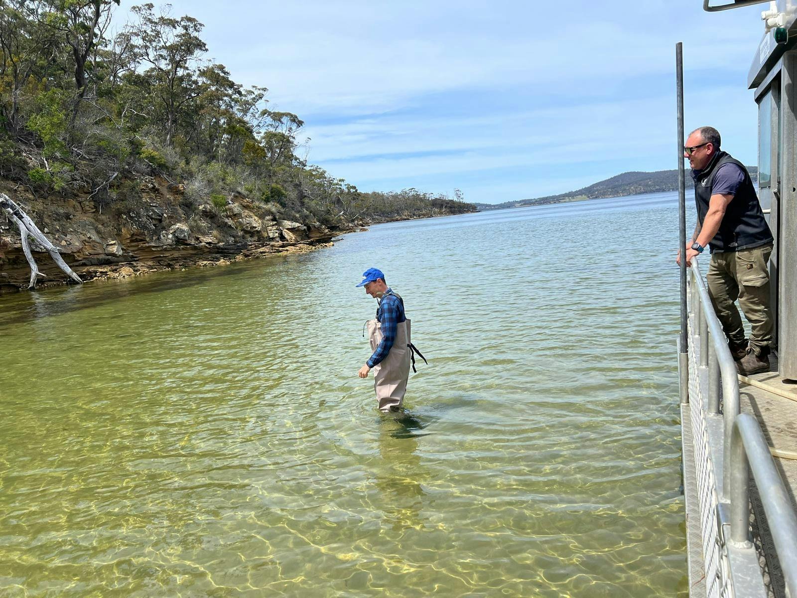 Person in waders stepping into clear water, observer watches from boat by forested shore