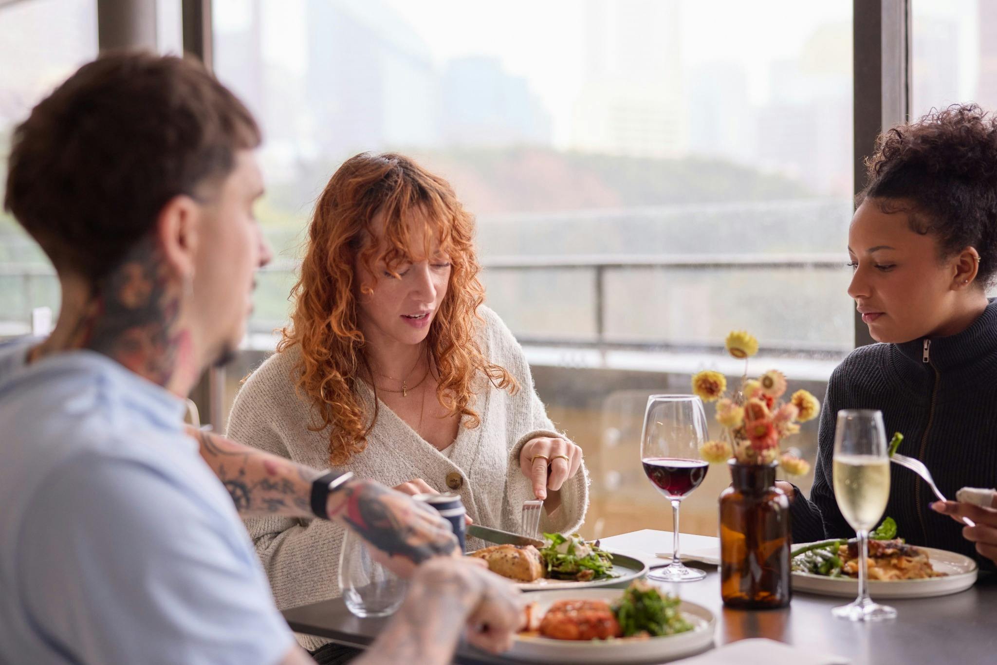 Three guests enjoying lunch and drinks in a light-filled restaurant with city views.