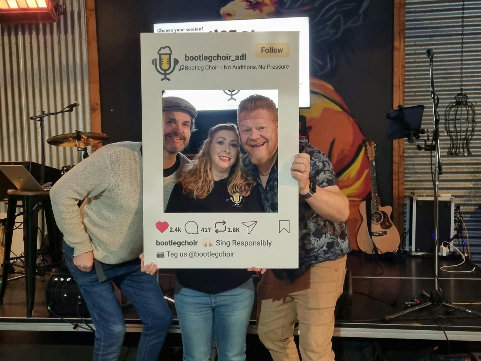 Three smiling adults pose behind a giant Instagram-style frame on a concert stage