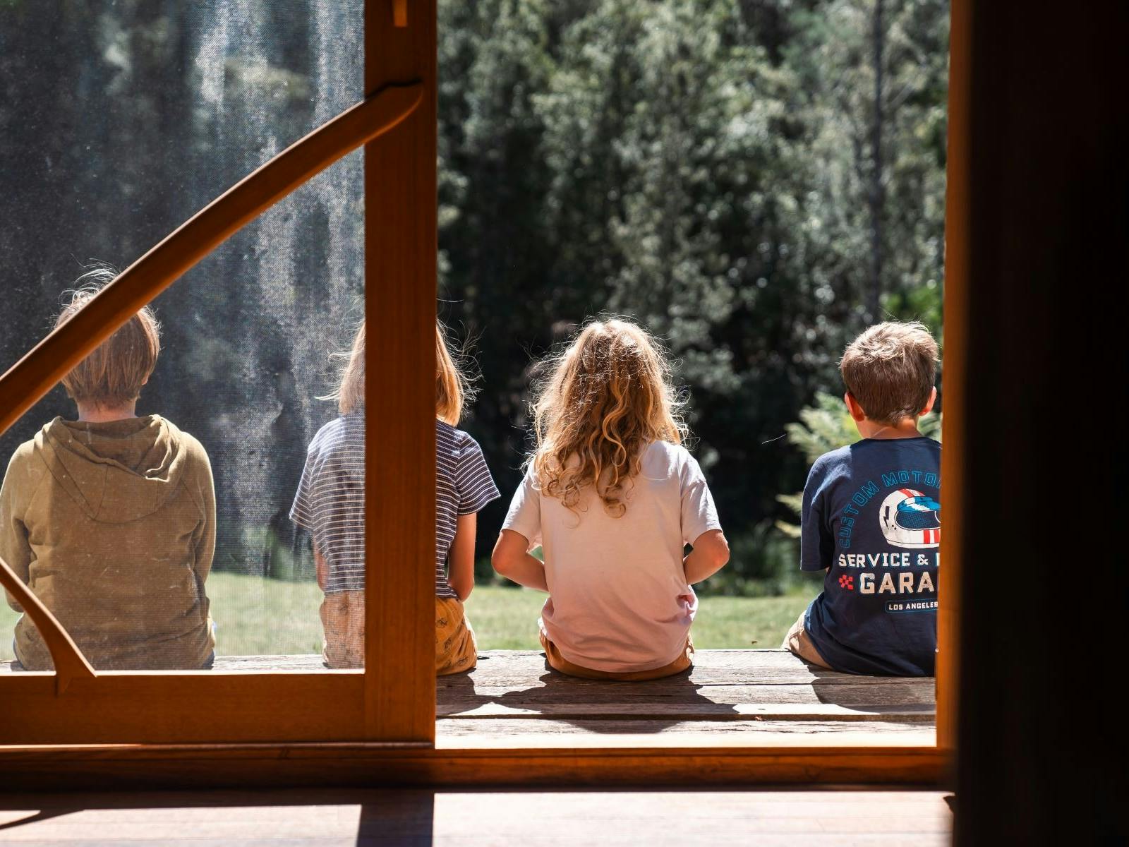 Four children sit on a sunny deck, viewed from inside through glass and timber doors.
