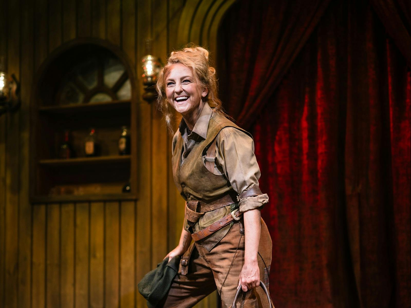 Blonde haired woman dressed in moleskins and vest holding a cap and whip in either hand
