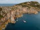 Aerial view of boat in ocean next to rocky cliffs on a peninsula with the ocean in the distance