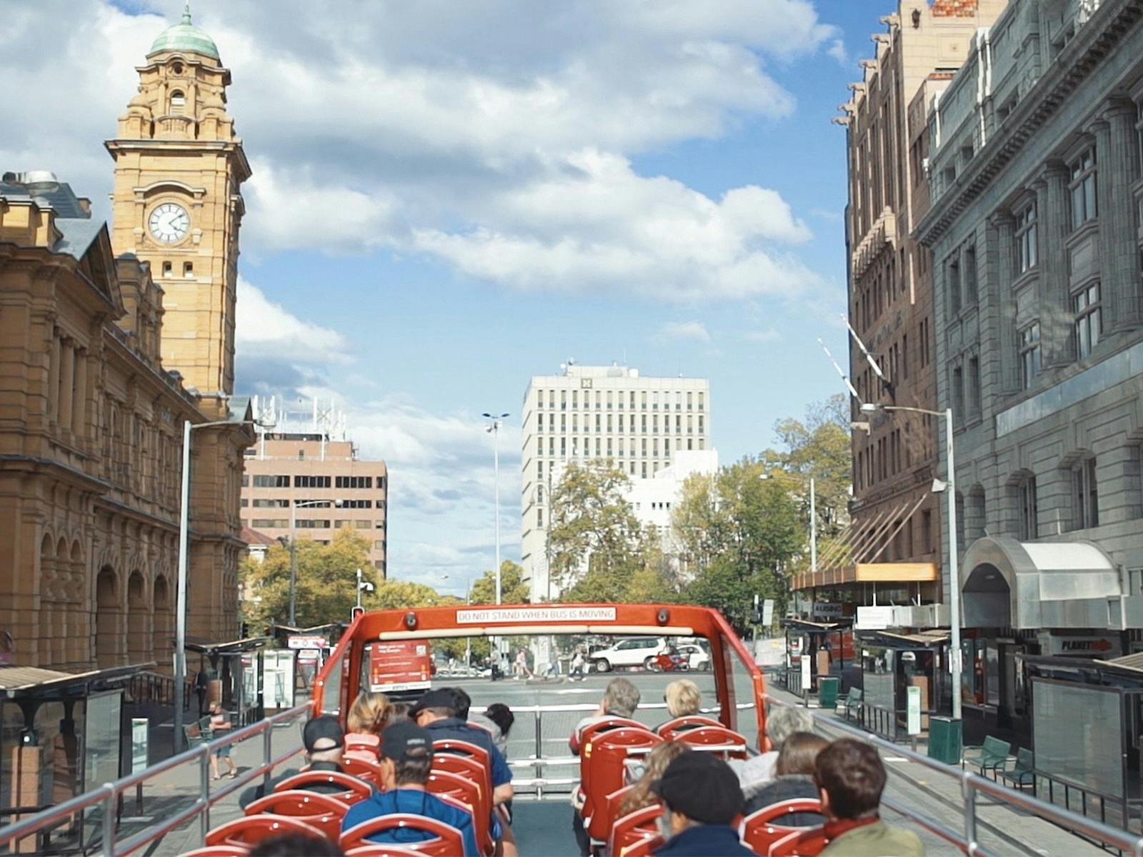 Passengers sitting on open-top double decker bus while it drives past city buildings.