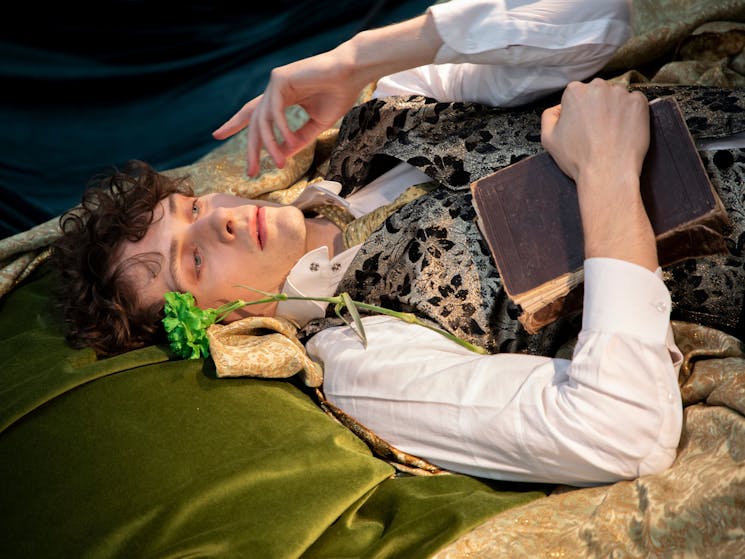 A dancer dressed as Oscar Wilde lying on a green bed and holding a book.