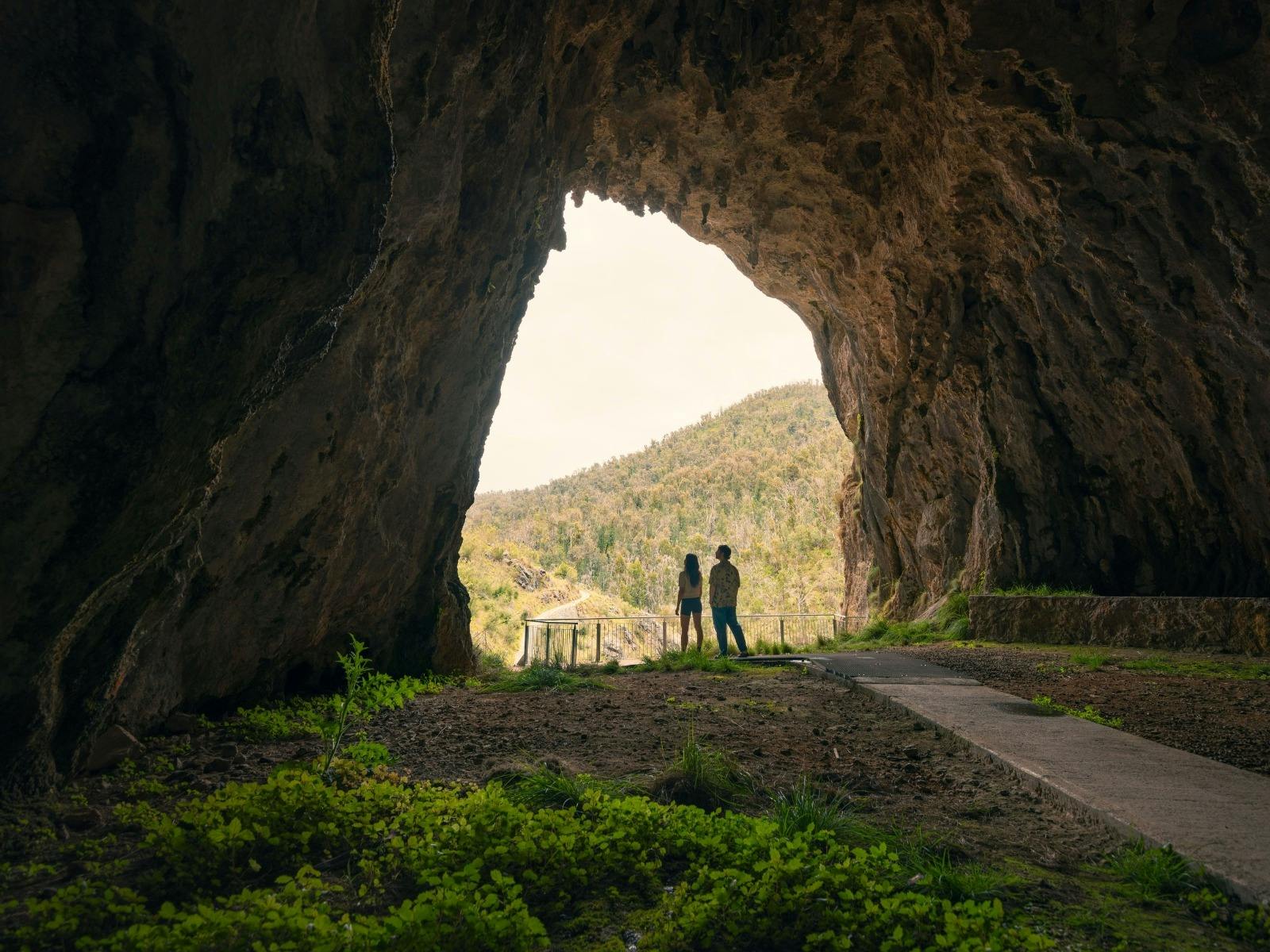 A couple seen from a distance at the entrance of a cave, with a bright sunny landscape ahead of them
