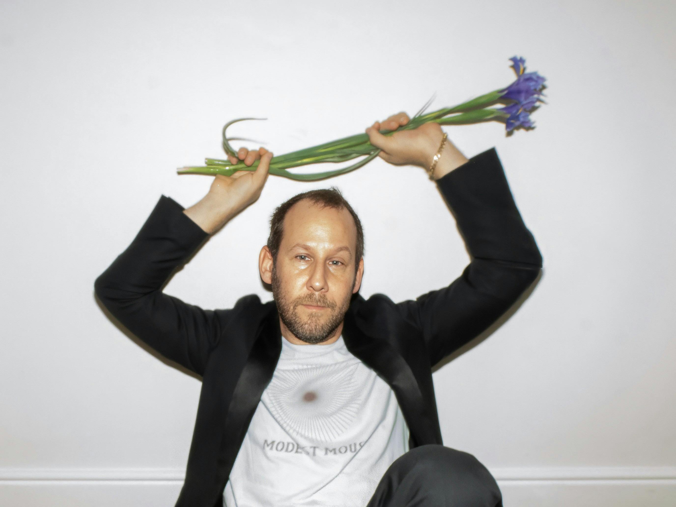 A man in a black blazer and white tee sits against a white wall, holding flowers above his head.