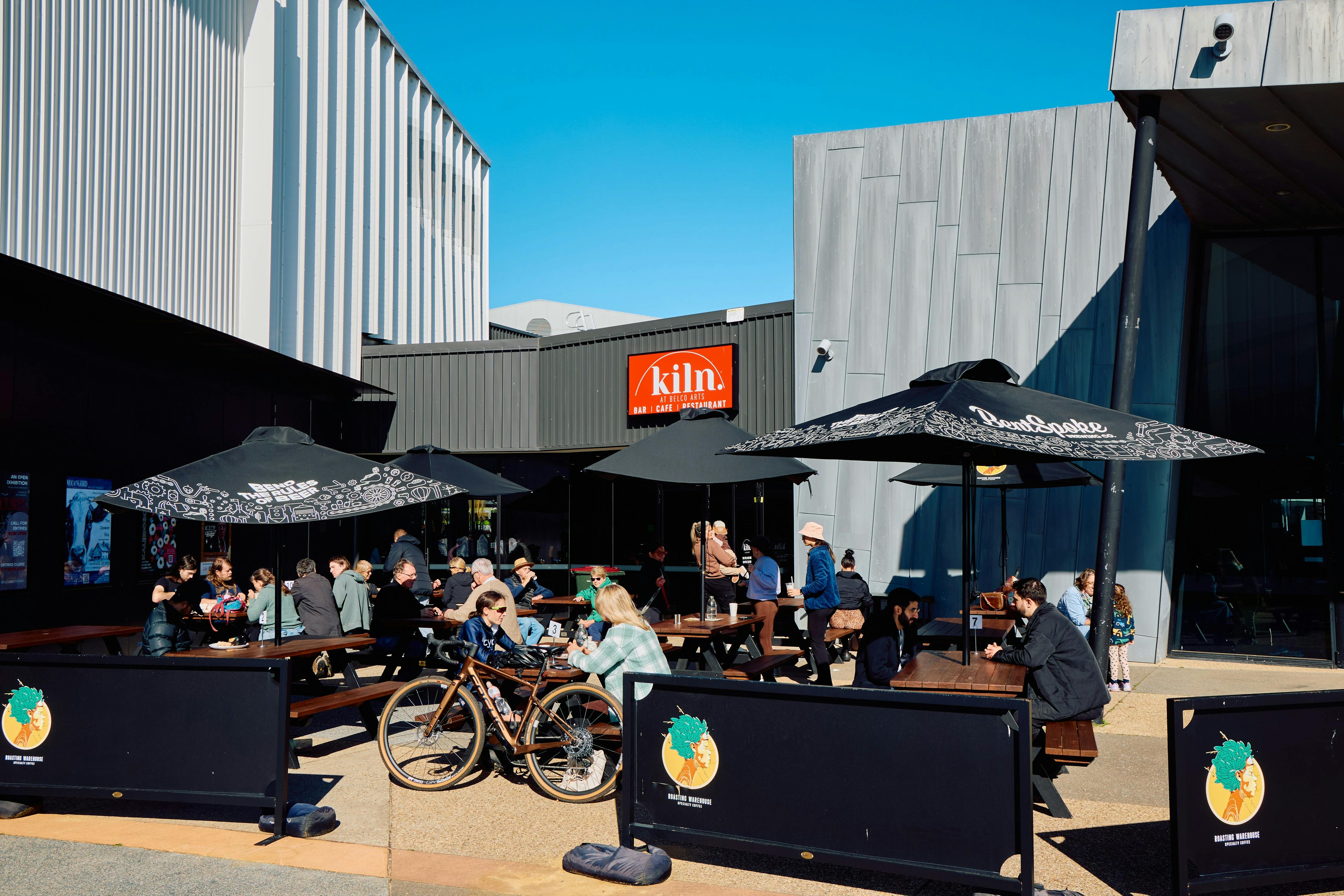 The outdoor terrace of Kiln café at Belco Arts. People eat and drink beneath parasols on a sunny day