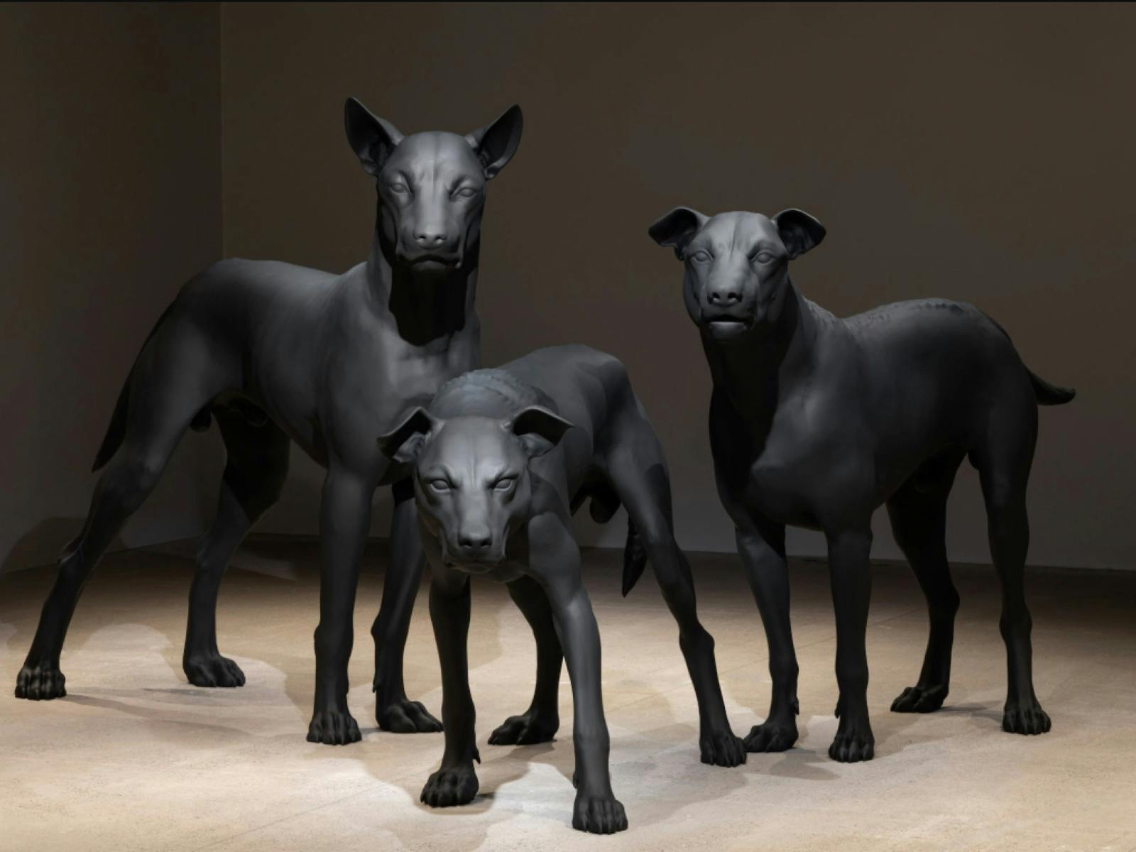 Three life-sized, matte black dog sculptures stand alert in a dimly lit gallery