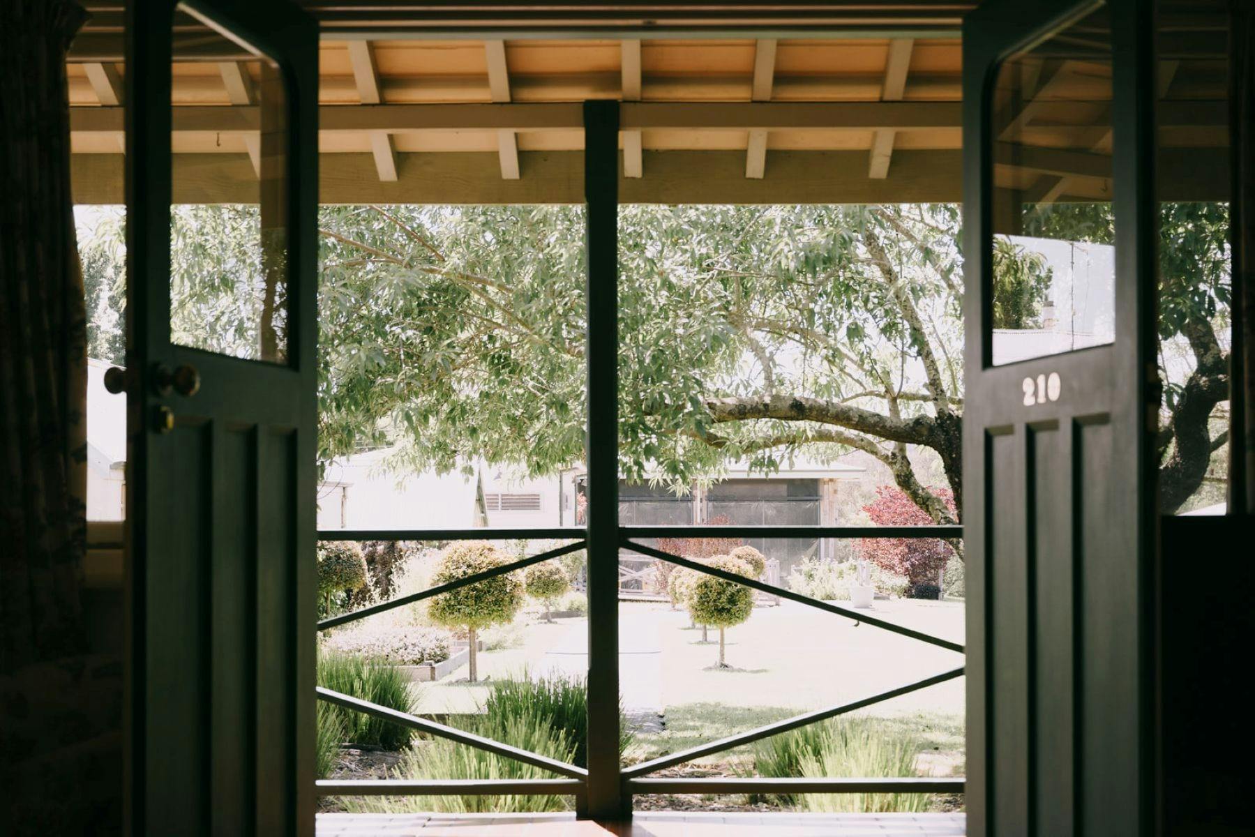View from an open doorway onto a garden with trees, manicured shrubs, and a covered porch railing.