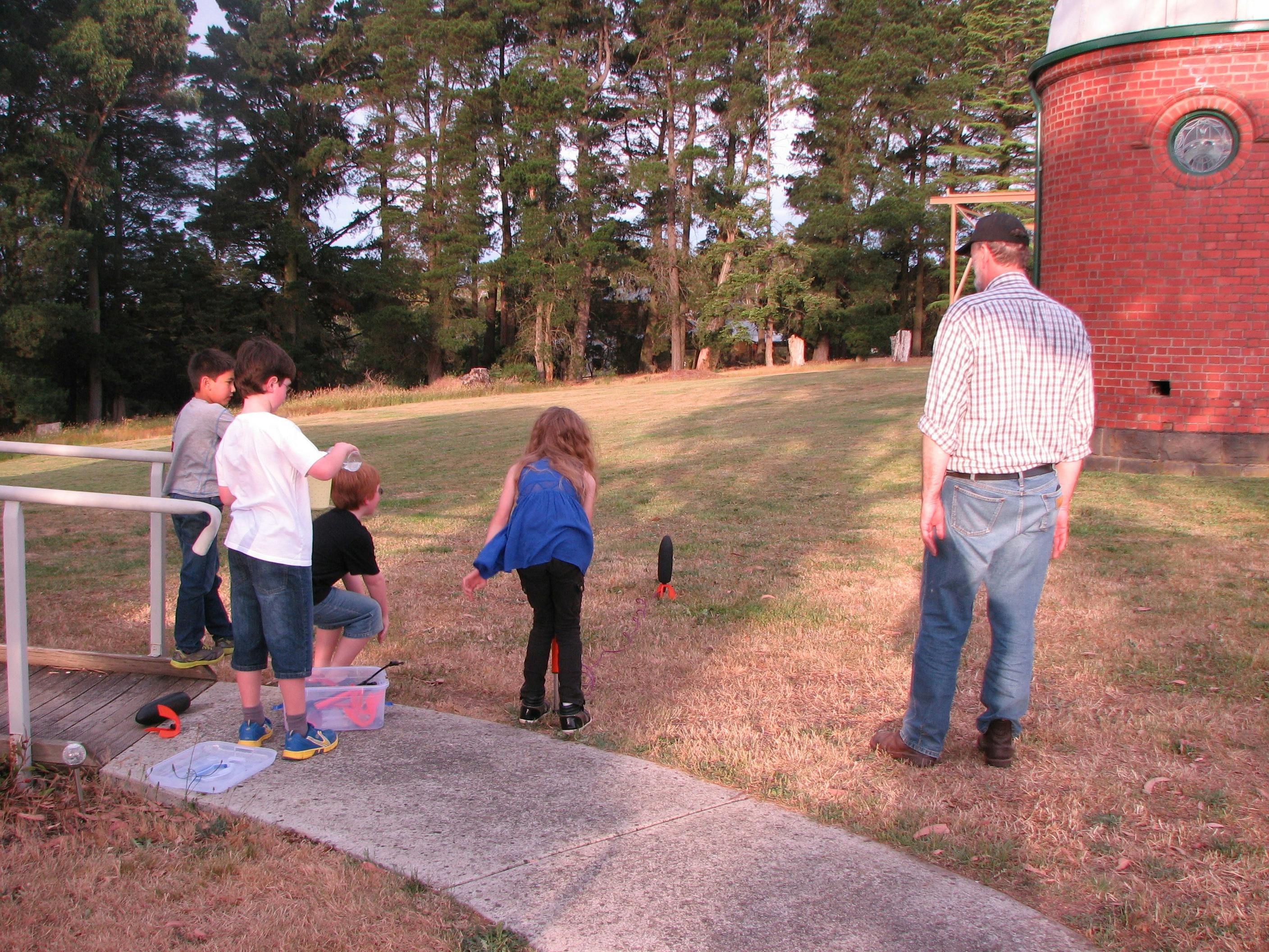 Children stand around a water rocket in front of a dome building.